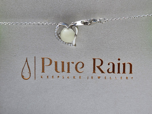 A heart-shaped cubic zirconia pendant necklace with a silver setting on an adjustable chain, presented against a textured grey background with the brand name 'Pure Rain Keepsakes'.